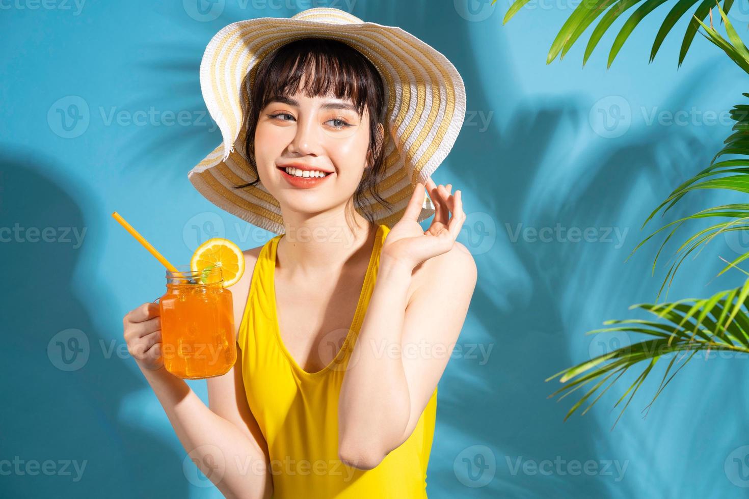 vacker asiatisk kvinna som bär gul jumpsuit på blå bakgrund och dricker juice, sommarbegrepp foto