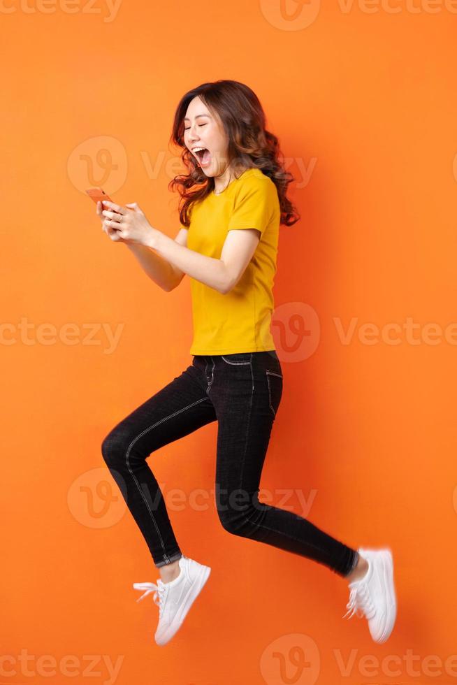 ung asiatisk kvinna som använder telefonen och hoppar på orange bakgrund foto