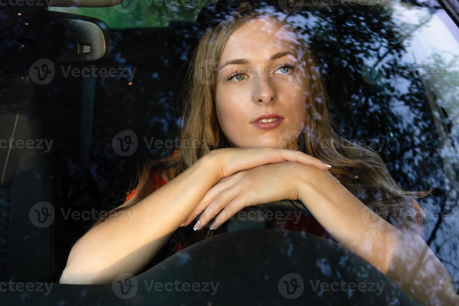 en ung, vacker kvinna med långt hår sitter vid ratten på bilen och tittar drömande genom bländningen på den främre vindrutan. foto