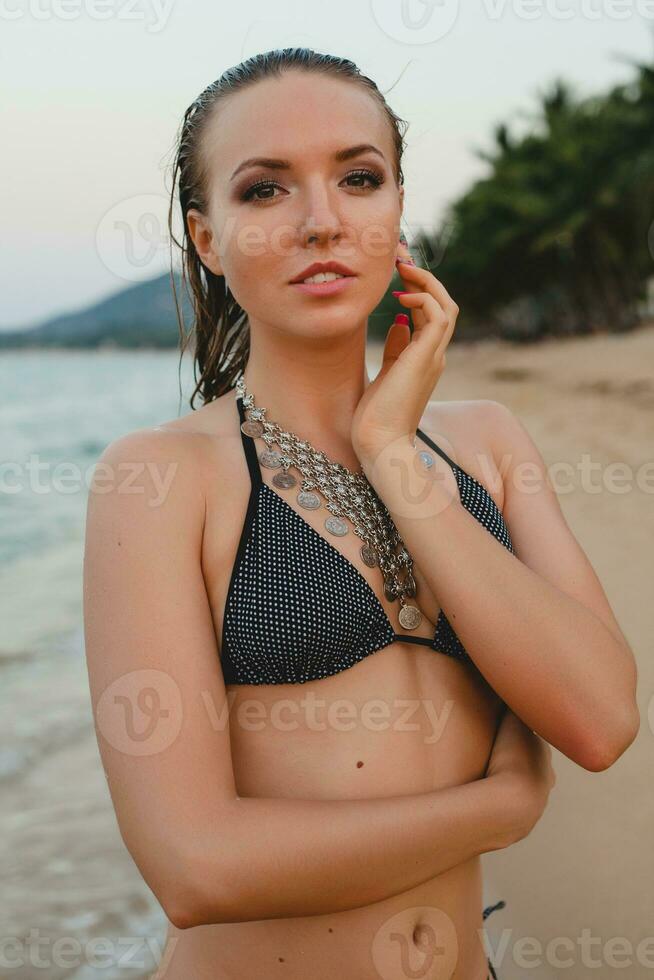 ung skön blond kvinna solbad på sand strand i bikini simning kostym, årgång halsband foto