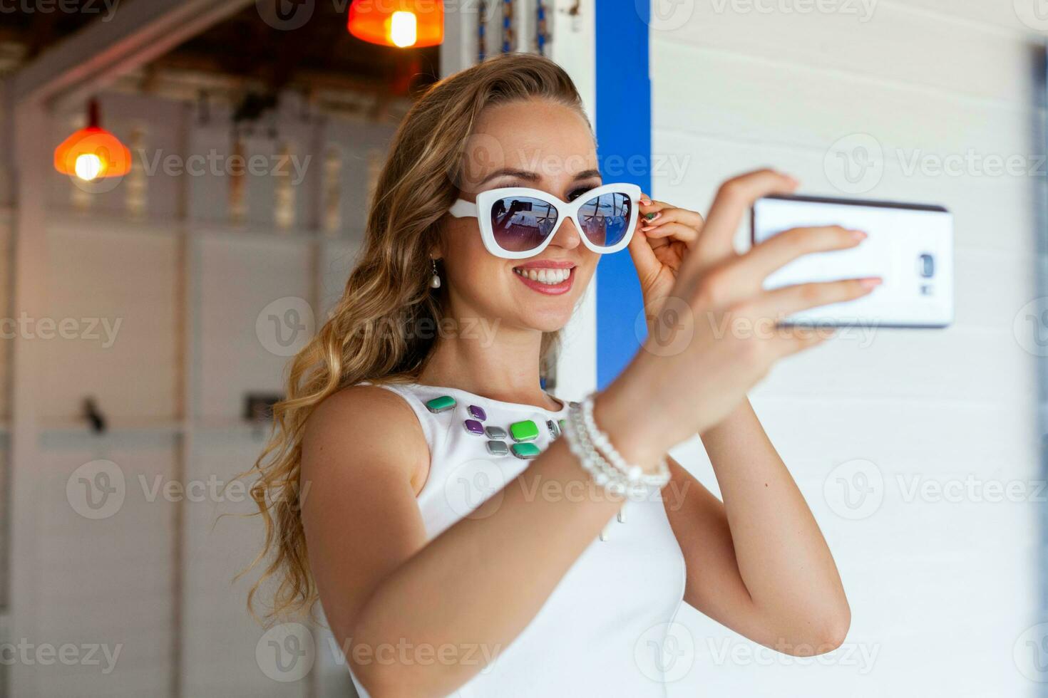 attraktiv kvinna i vit klänning i sommar Kafé i solglasögon använder sig av telefon tar Foto