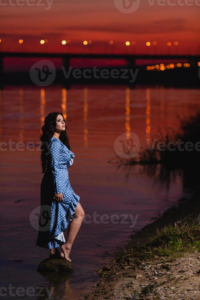 vacker ung flicka med långt mörkt vågigt hår som står vid stranden av floden foto