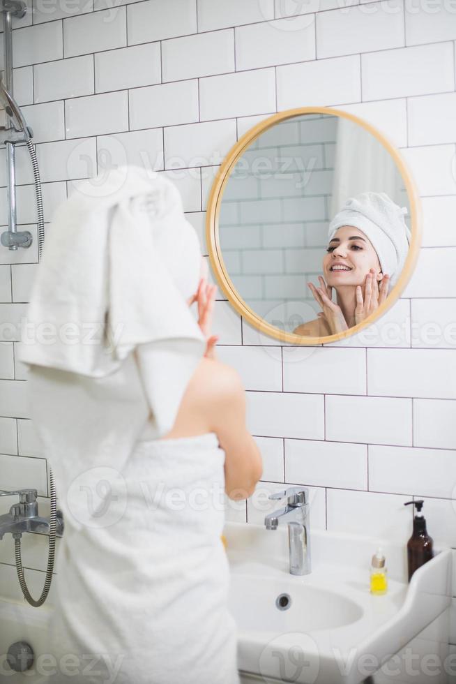 ung kvinna i vit handduk applicera fuktgivande olja på ansiktshuden. foto