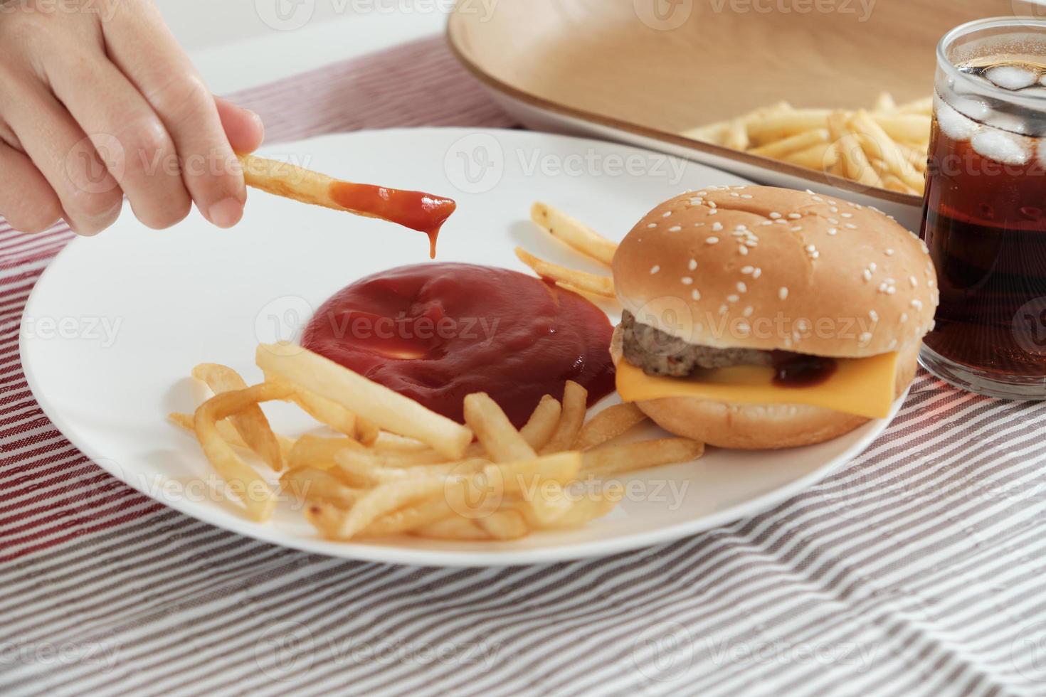 närbild, selektiv fokus. mans hand åt fransk stekt, doppade ketchup på en vit tallrik på röd bordsduk med hamburgare och cola. att äta skräpmat eller snabbmat till lunch är ohälsosamt. foto