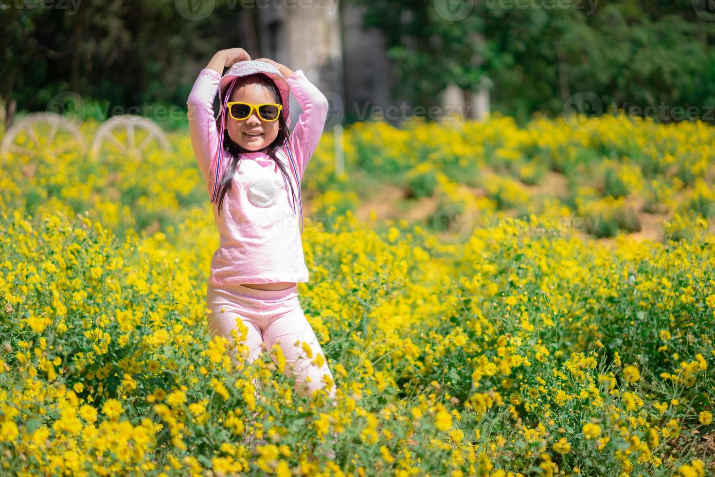 liten asiatisk tjej i rosa klänning bär hatt och solglasögon som står i gul blommaträdgård foto