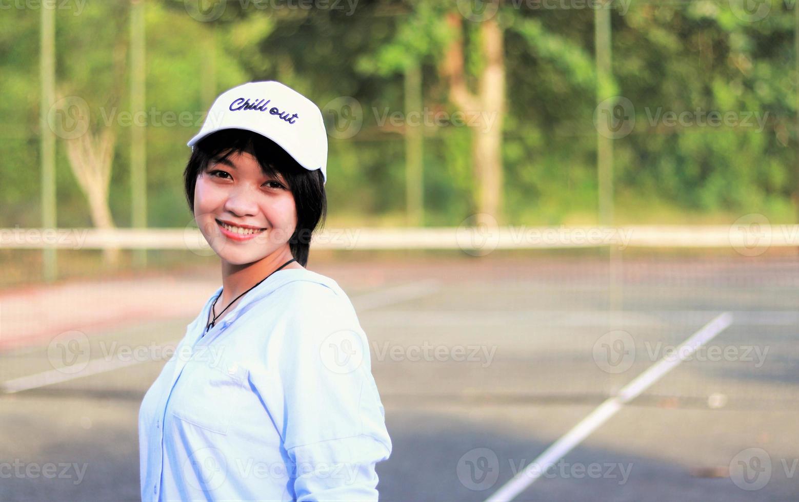 vacker asiatisk kvinna med kort hår, hatt och ler bredt på tennisbanan foto