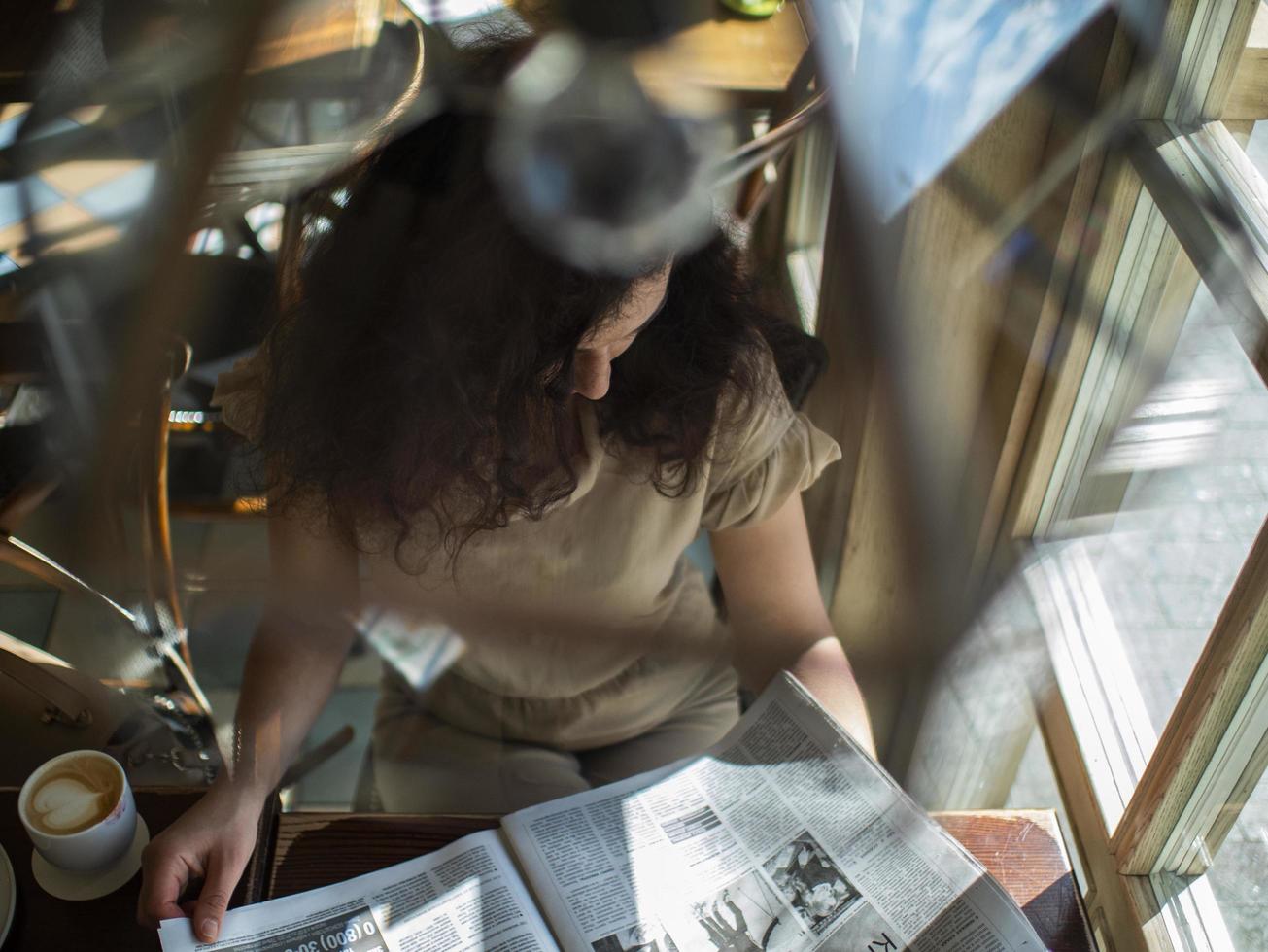 en ung flicka med lockigt hår sitter vid ett bord på ett kafé och läser en tidning foto