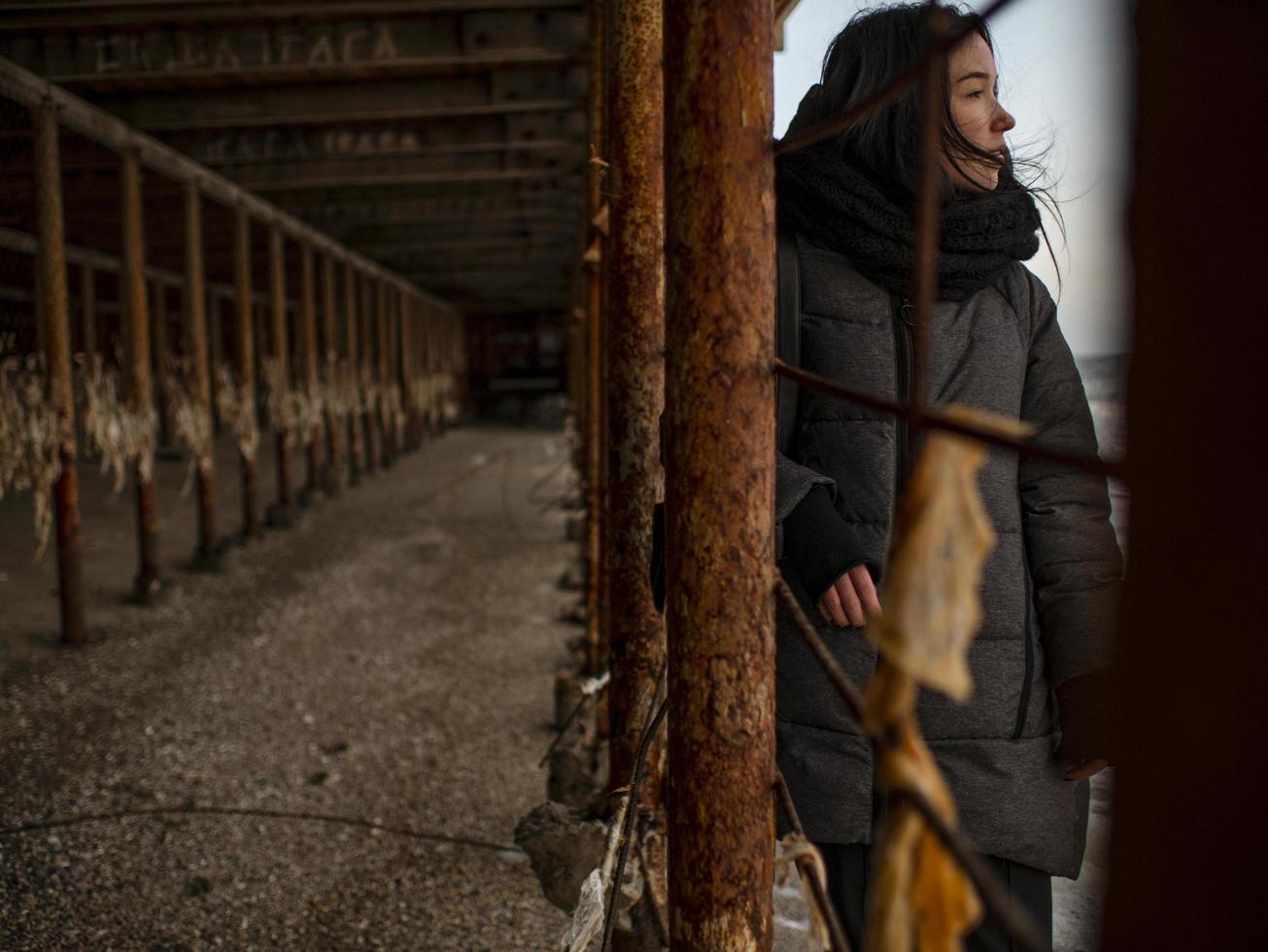 ung flicka i en kappa står nära metall rostiga strukturer nära havet kusten foto