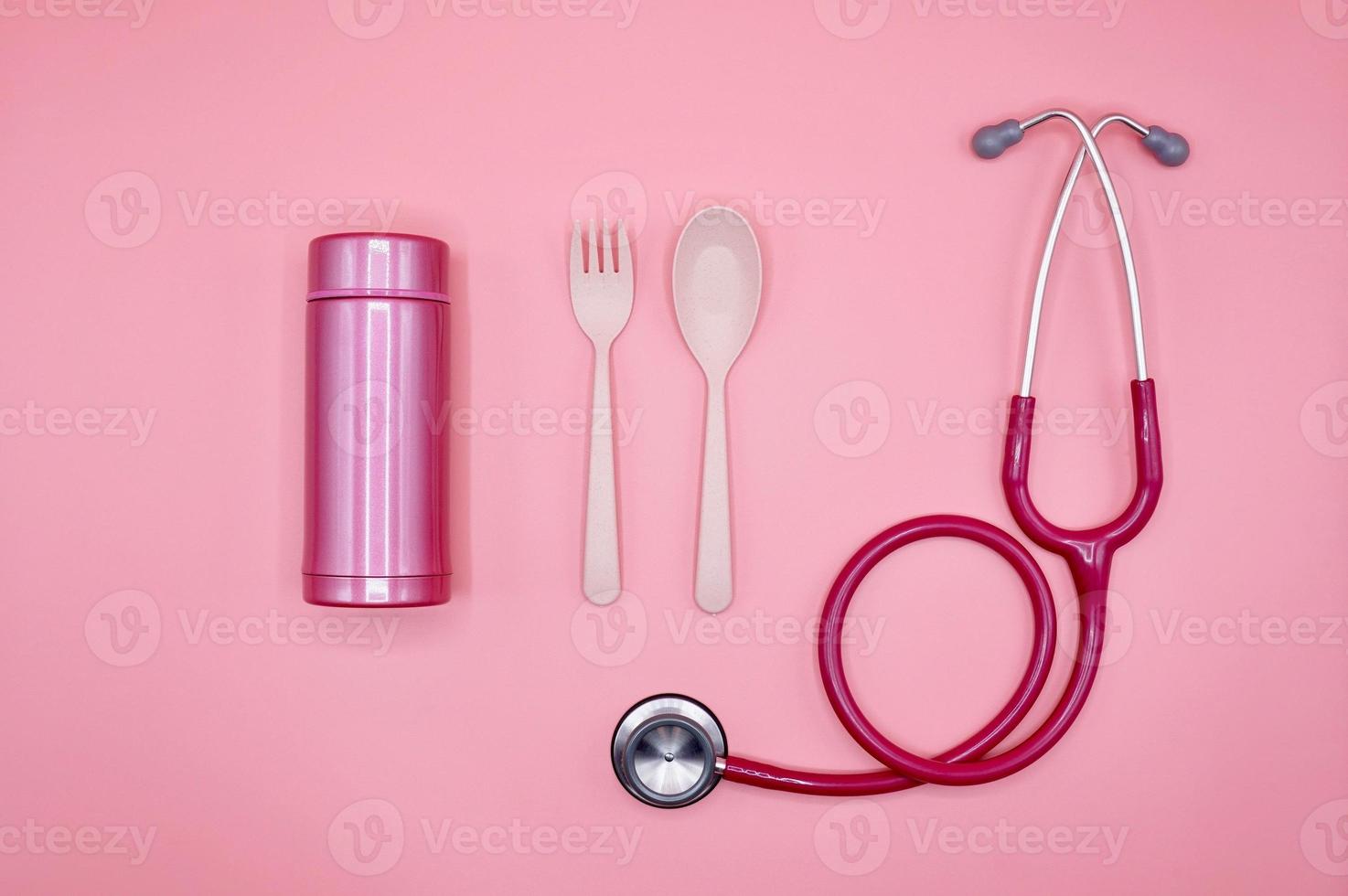 ett rosa stetoskop, sked, gaffel och termosflaska på den rosa bakgrunden, platt layout foto