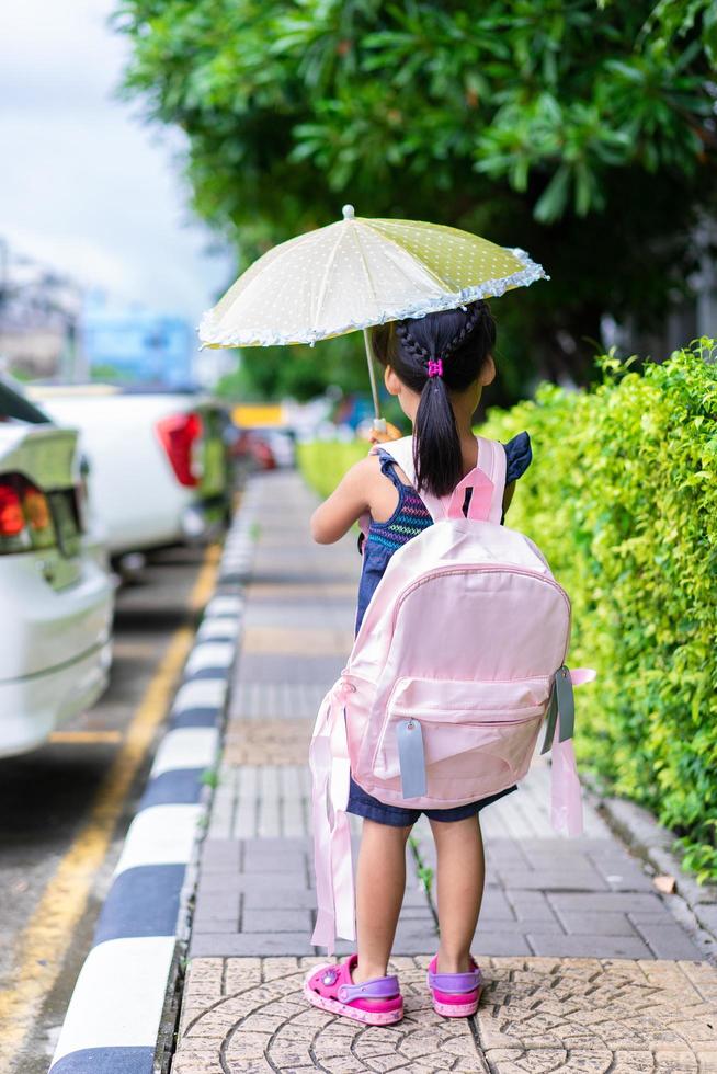 liten asiatisk tjej med paraply och ryggsäck promenader i parken redo tillbaka till skolan i regnar dag foto