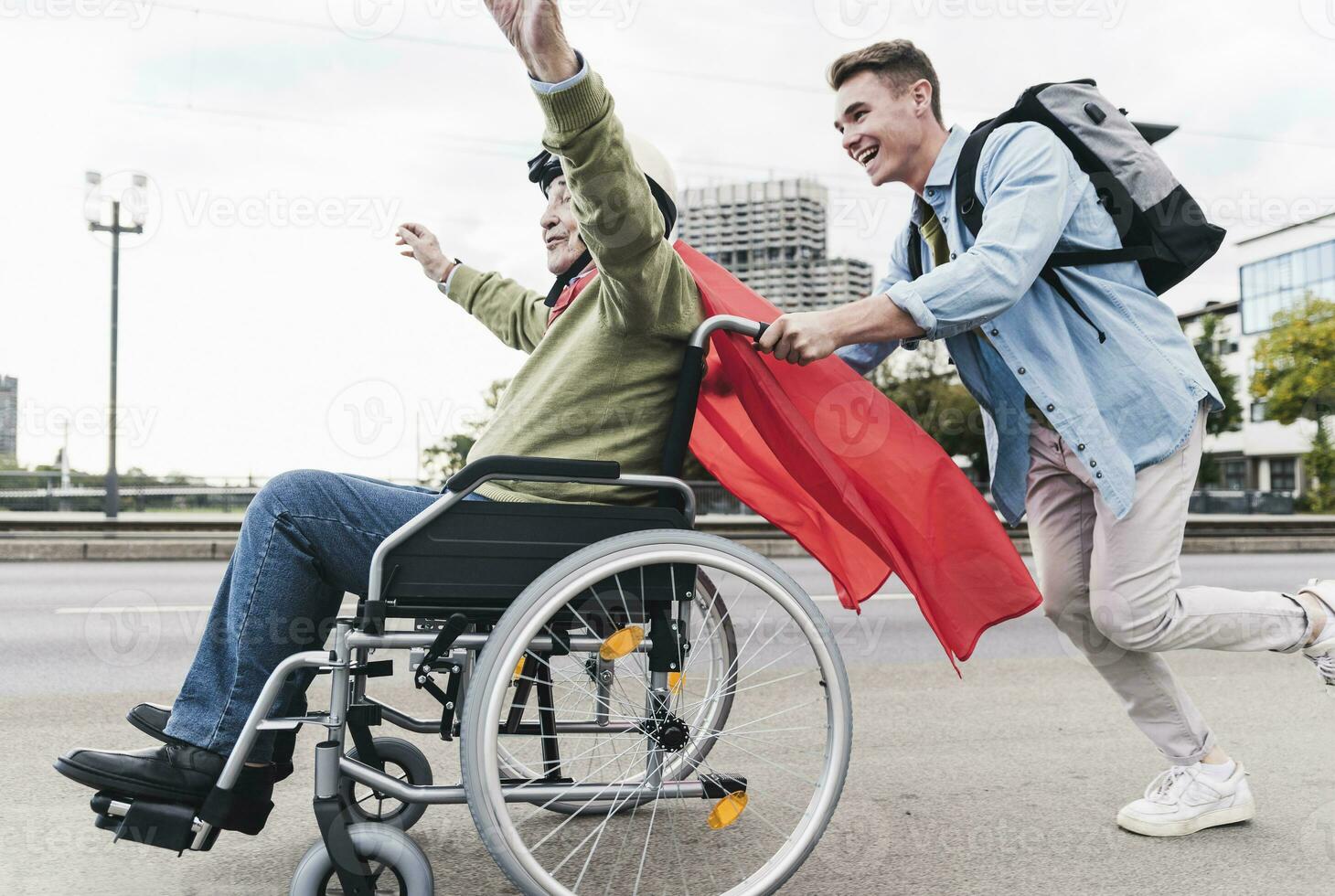 ung man tryckande senior man Sammanträde i en rullstol klädd upp som superhjälte foto