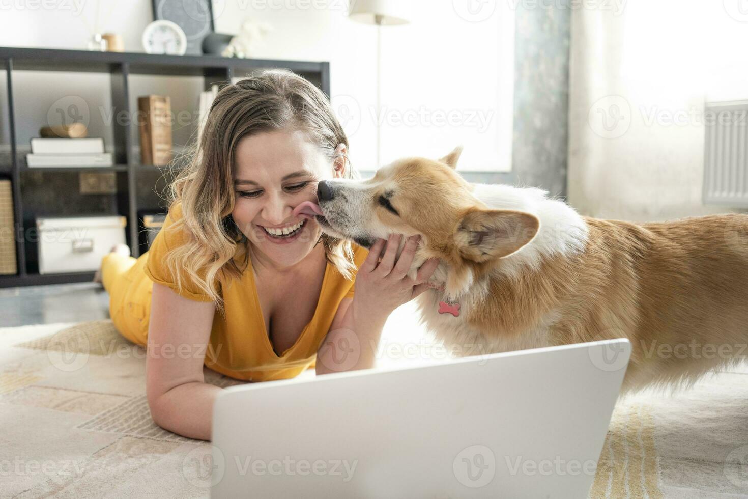 Lycklig kvinna använder sig av bärbar dator i levande rum på Hem med hund slick över henne ansikte foto