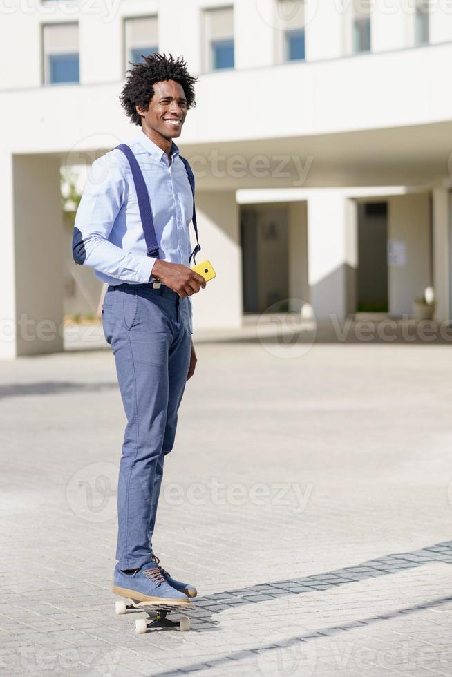 svart affärsman på en skateboard som håller en smartphone utomhus. foto