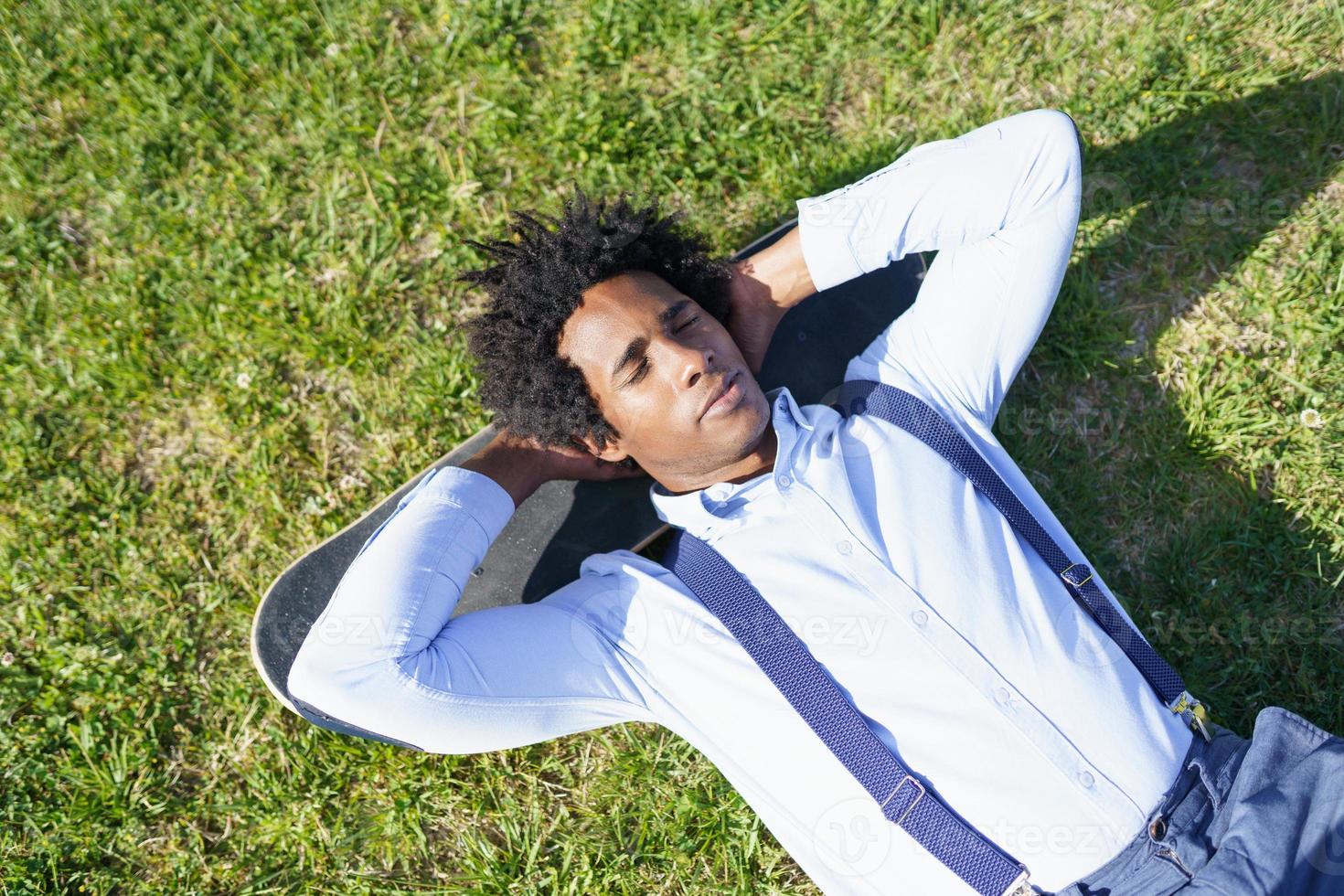 svart affärsman vilar liggande på gräset med sin skateboard foto