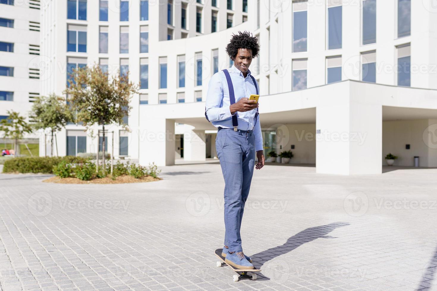 svart affärsman på en skateboard tittar på sin smartphone utomhus. foto