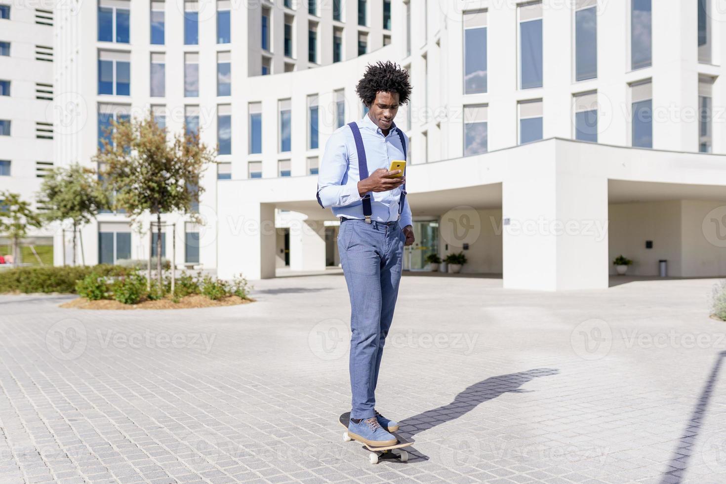 svart affärsman på en skateboard tittar på sin smartphone utomhus. foto