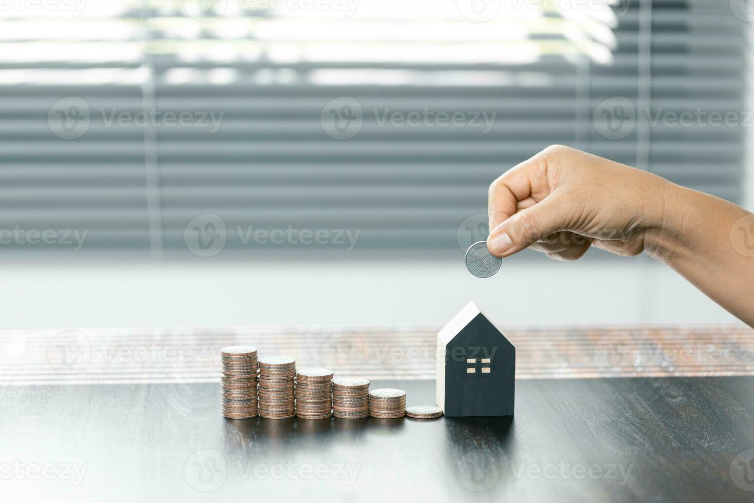 sparande pengar för hus och verklig egendom. kvinna hand innehav en mynt med stack mynt och hus modell på tabell. foto