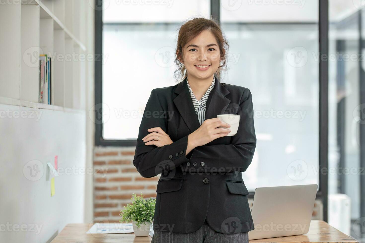 porträtt, självsäker leende asiatisk affärskvinna i kostym stående i kontor med vapen vikta och innehav kaffe kopp. foto