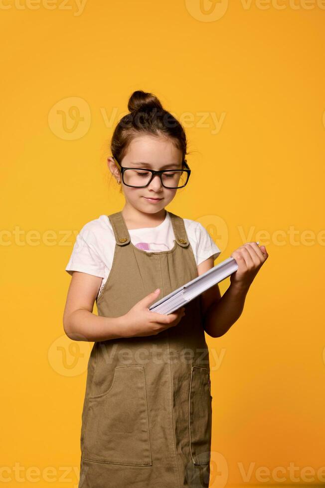 vertikal studio porträtt av självsäker allvarlig liten unge flicka 6 år gammal, innehav lärobok, isolerat orange bakgrund foto