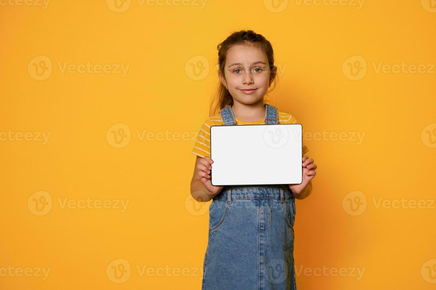 förtjusande barn flicka som visar digital läsplatta med vit tömma tom skärm, leende ser på kamera, isolerat bakgrund foto