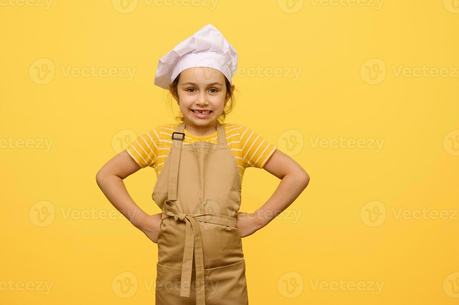 busig barn flicka klädd som konditor i förkläde och vit kockens keps, sätta händer på midja, ser på kamera foto