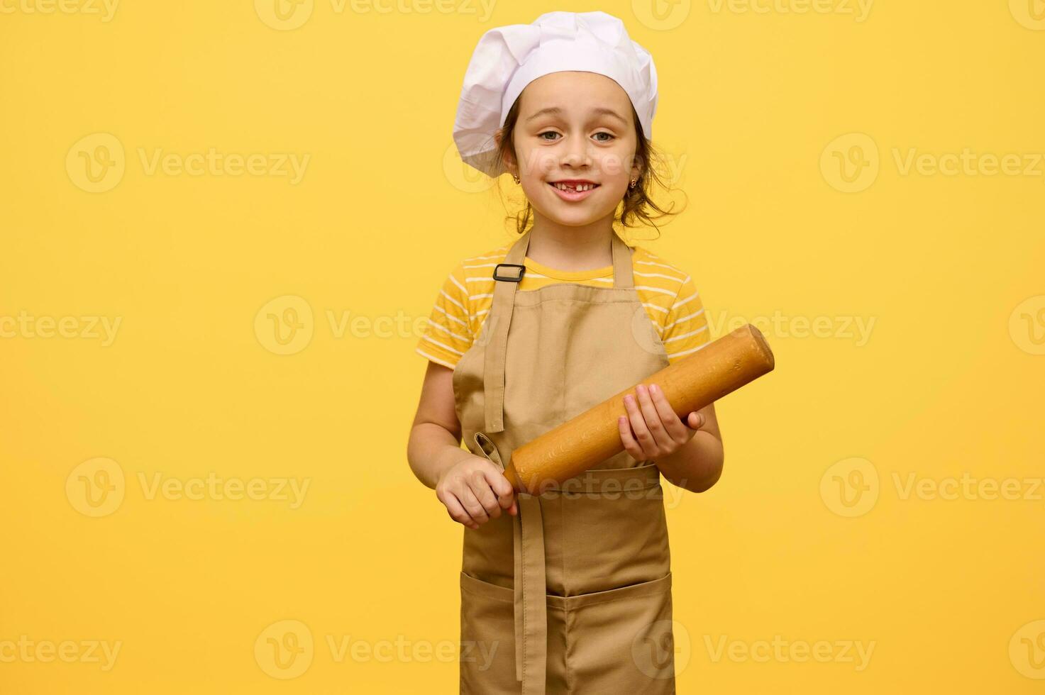 Lycklig liten förskola flicka med rullande stift, klädd som kock bakverk, leende ser på kamera, gul bakgrund foto