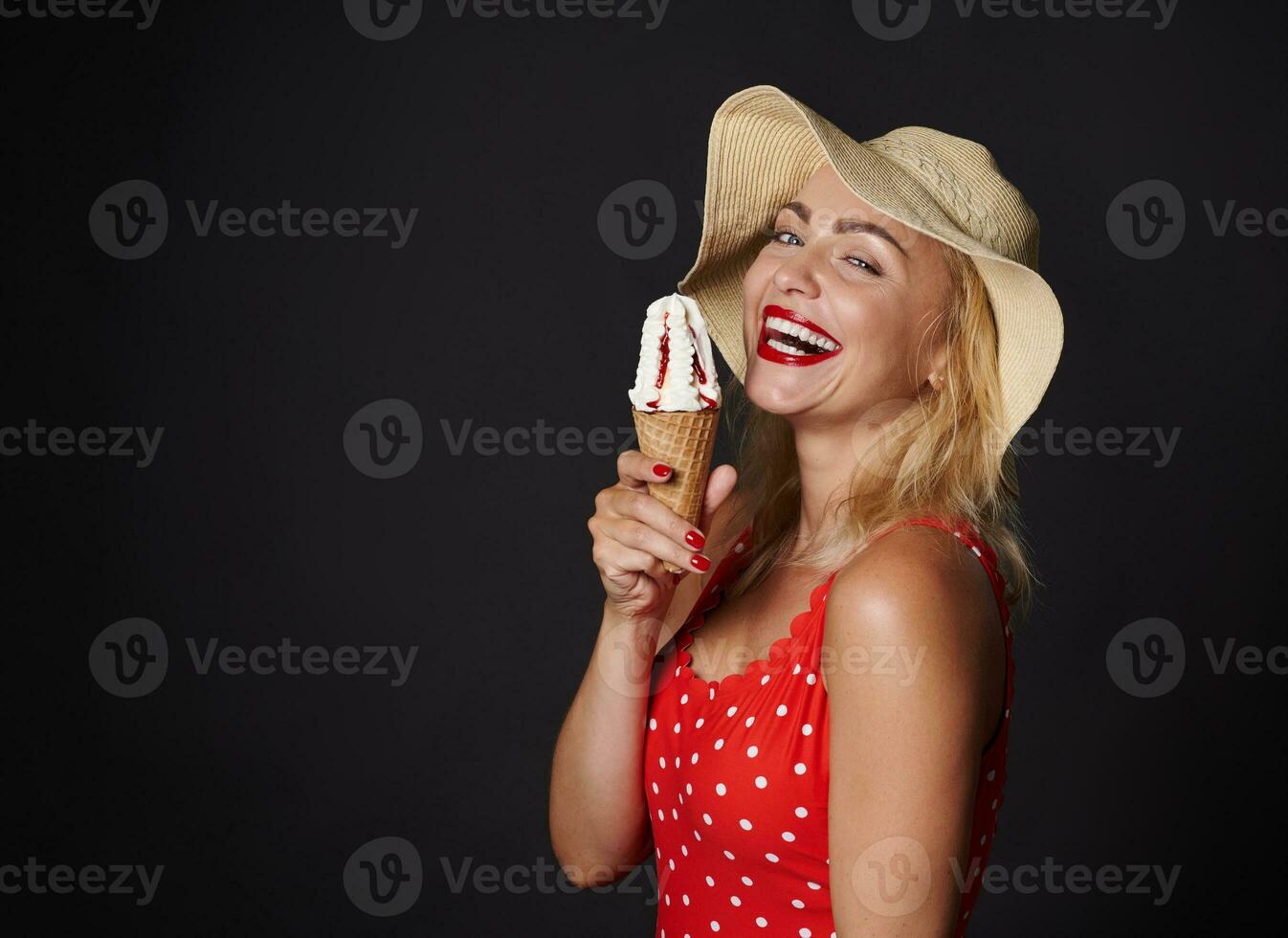 attraktiv blond europeisk kvinna i röd baddräkt med polka prickar och sugrör hatt innehar gott kornett is grädde, visar och ler sött med skön toothy leende ser på kamera över svart bakgrund foto