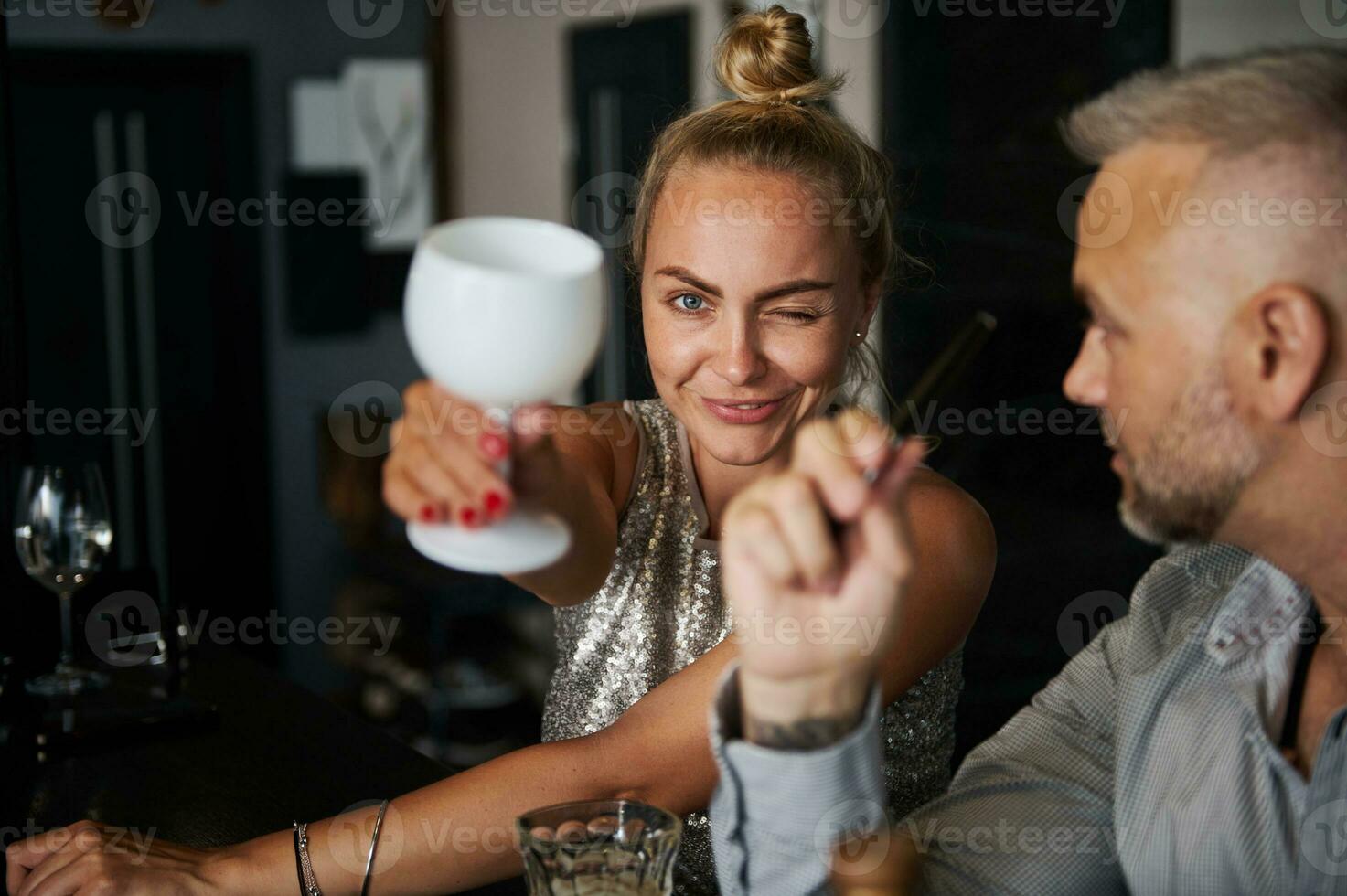 fokus på en glad mitten åldrig attraktiv blond kvinna, blinkning och leende som visar en vit dricka glas i henne utsträckt hand till de kamera, Sammanträde stänga till henne partner på ny år fest foto