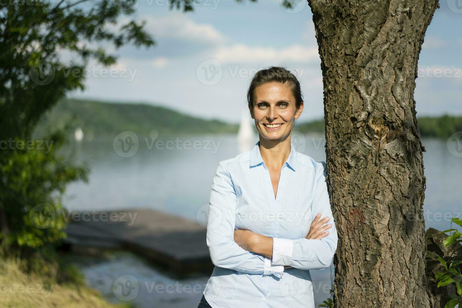 porträtt av leende kvinna lutande mot en träd på en sjö foto