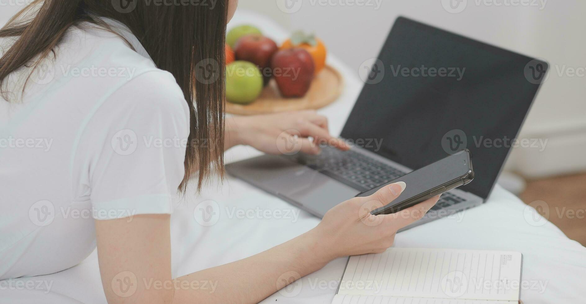 effektiv ung tusenåriga flicka Sammanträde på en säng i de morgon, användningar bärbar dator dator och äter croissanter och drycker kaffe för frukost foto