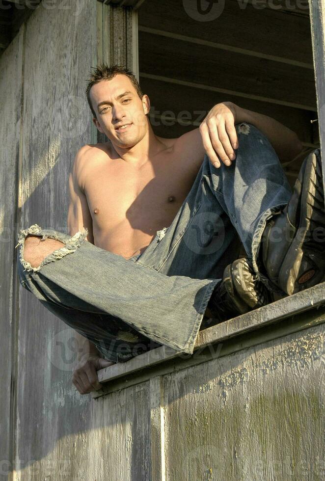 varm manlig modell sitter i en ladugård fönster med hans skjorta av och muskulös kropp visar av på vilket sätt sexig han är. foto