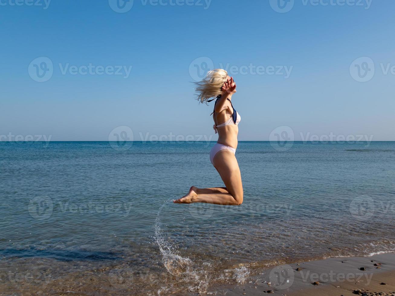blond flicka i bikini som hoppar upp med böjda ben, vattenstänk foto