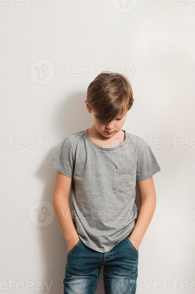 söt pojke som böjer huvudet och lutar sig mot den vita väggen foto
