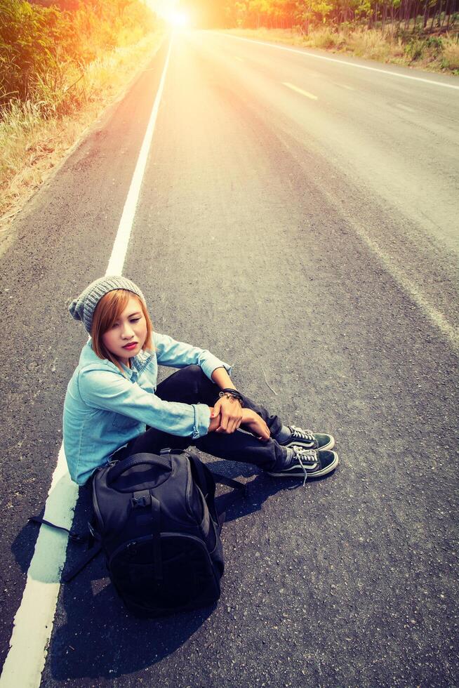 ung kvinna liftar bärande ryggsäck sitter på vägen foto