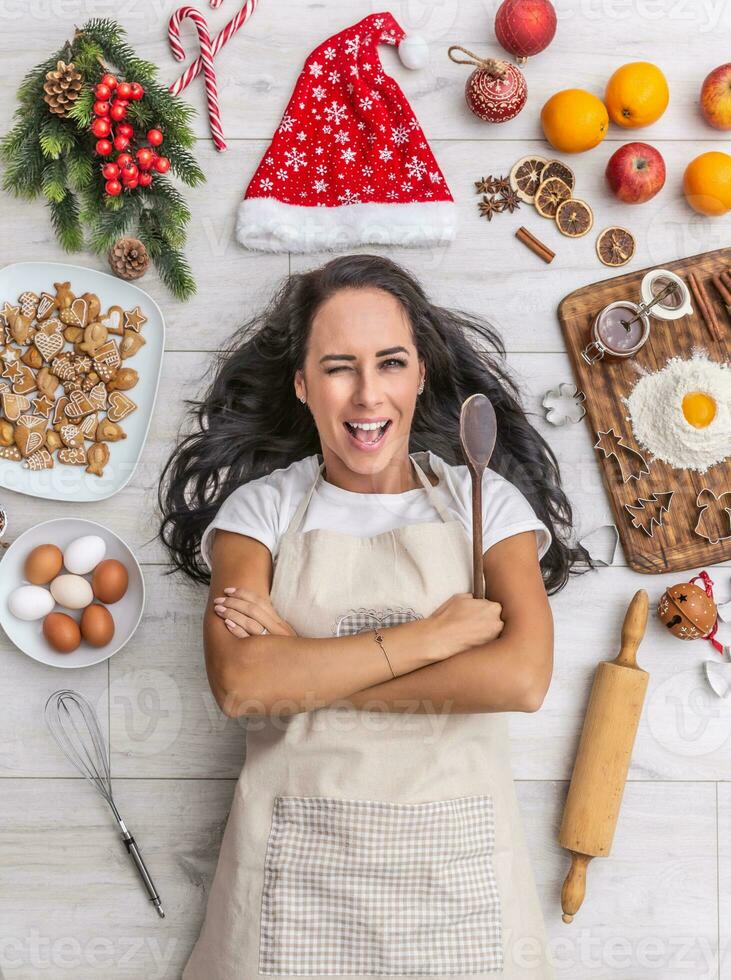 skön mörk håriga laga mat blinkning om och på de jord, innehav de trä- sked och varelse omgiven förbi pepparkakor, ägg, mjöl, jul hatt och frukt foto