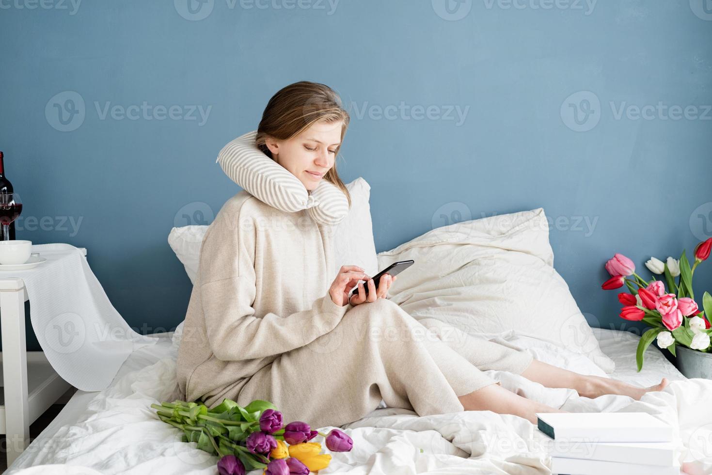 kvinna som sitter på sängen i pyjamas och skriver ett meddelande i telefonen foto