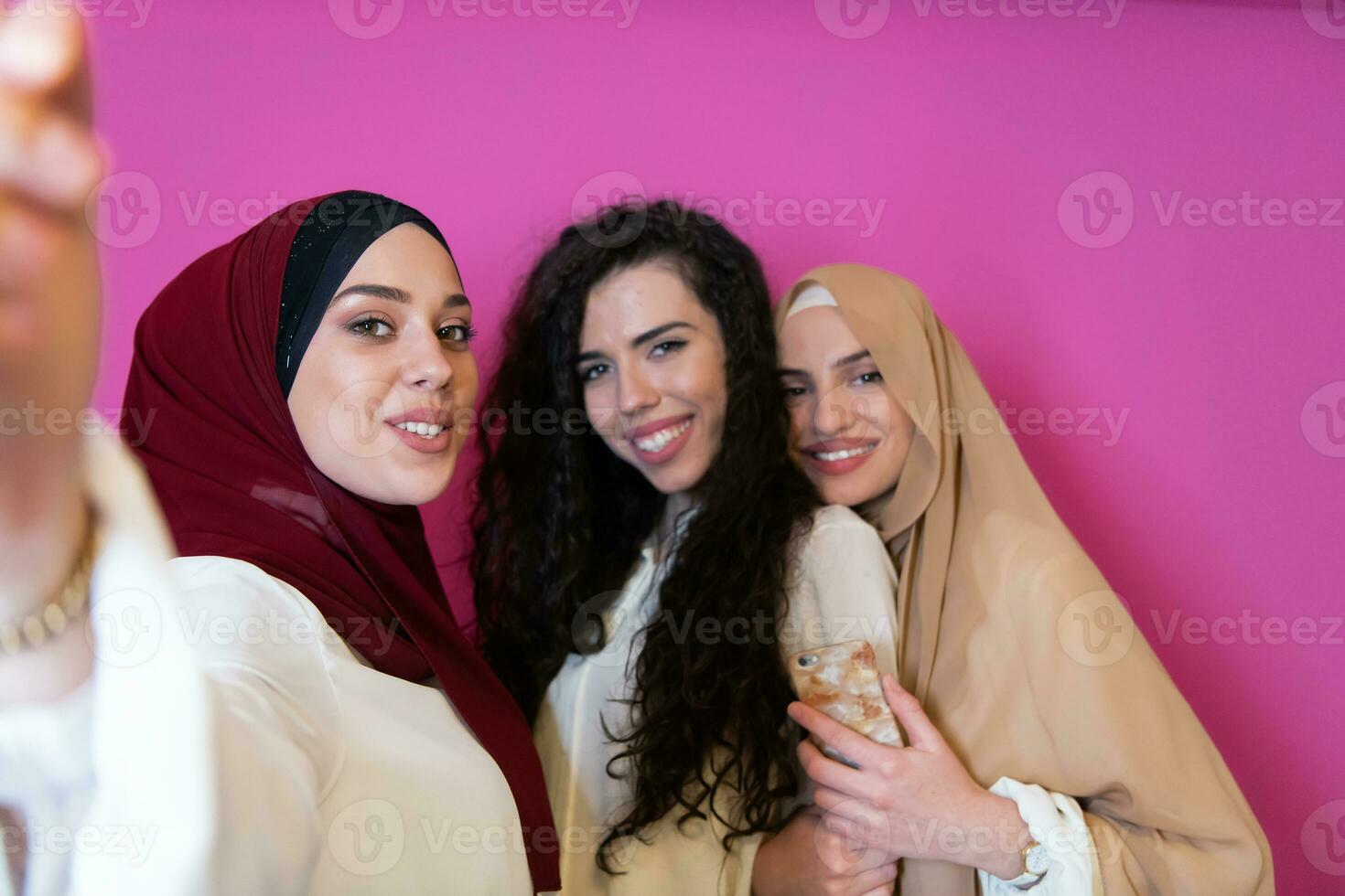 grupp av skön muslim kvinnor två av dem i modern klänning med hijab använder sig av mobil telefon medan tar selfie bild isolerat på rosa bakgrund representerar modern islam mode teknologi foto