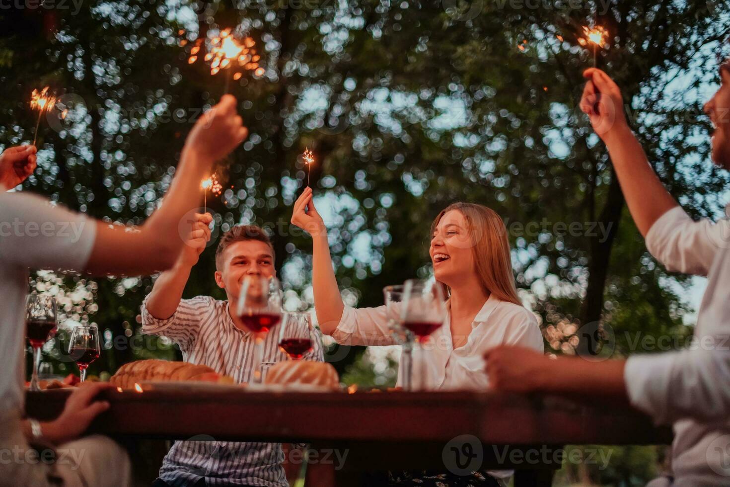 grupp av Lycklig vänner fira Semester semester använder sig av sprinklers och dricka röd vin medan har picknick franska middag fest utomhus- nära de flod på skön sommar kväll i natur foto