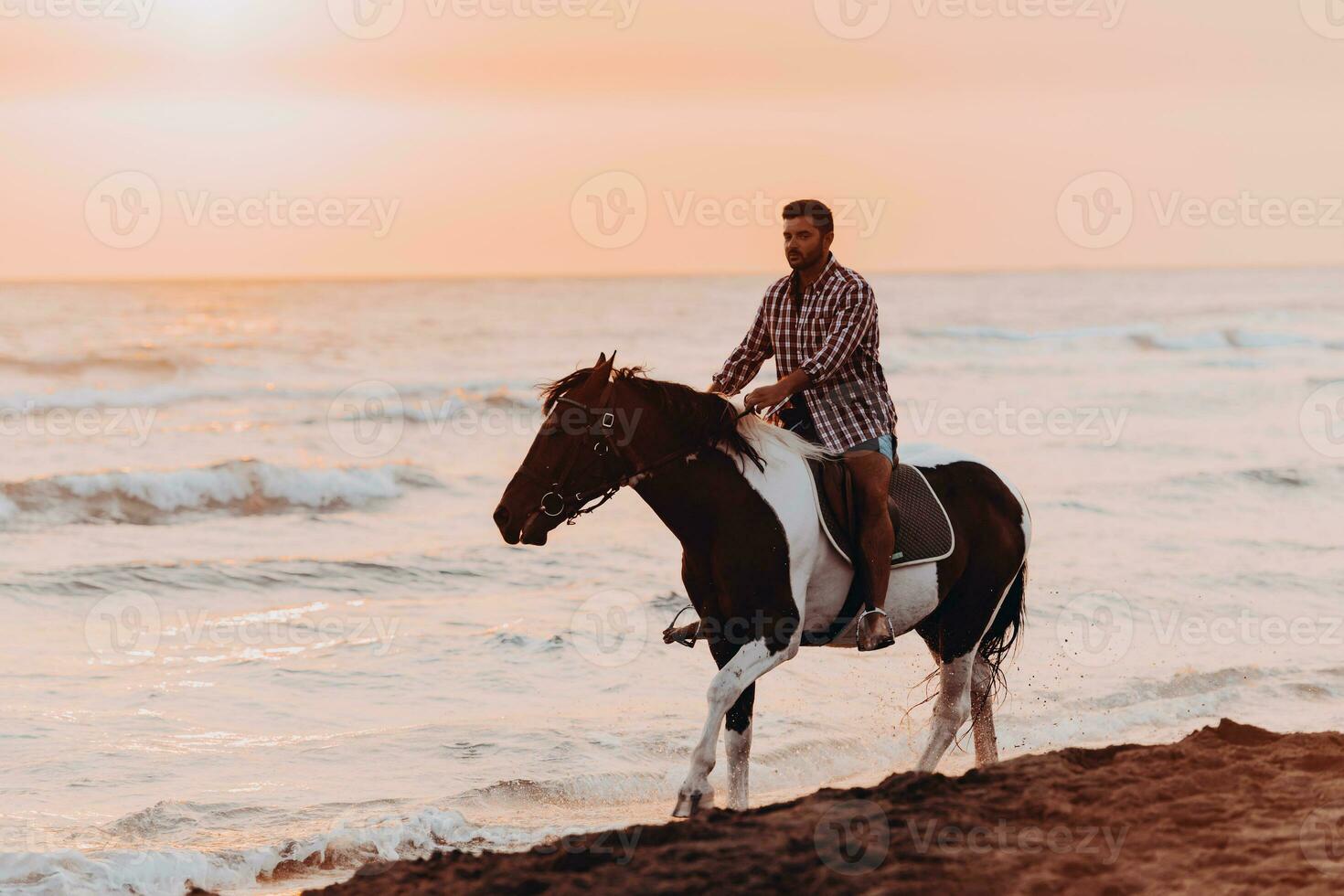 en modern man i sommar kläder åtnjuter ridning en häst på en skön sandig strand på solnedgång. selektiv fokus foto