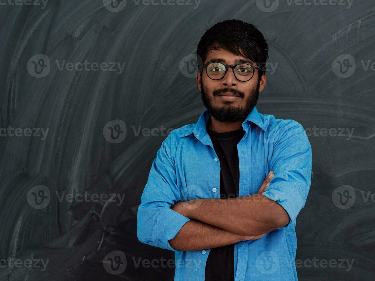 en ung indisk studerande i en blå skjorta med glasögon Framställ med hans vapen korsade i främre av de skola svarta tavlan foto