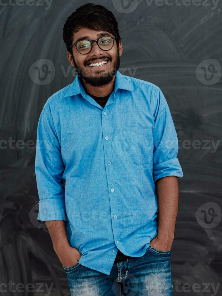 indisk leende ung studerande i blå skjorta och glasögon Framställ på skola svarta tavlan bakgrund foto