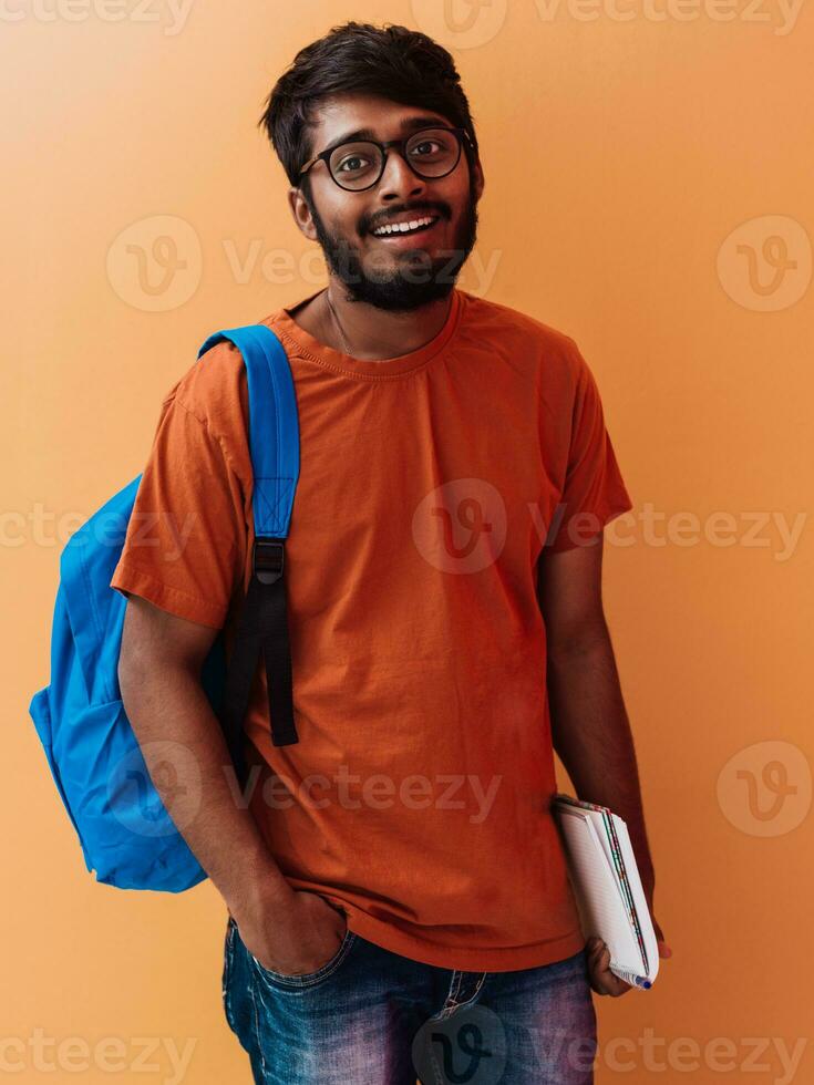 indisk studerande med blå ryggsäck, glasögon och anteckningsbok Framställ på orange bakgrund. de begrepp av utbildning och skolutbildning. tid till gå tillbaka till skola foto