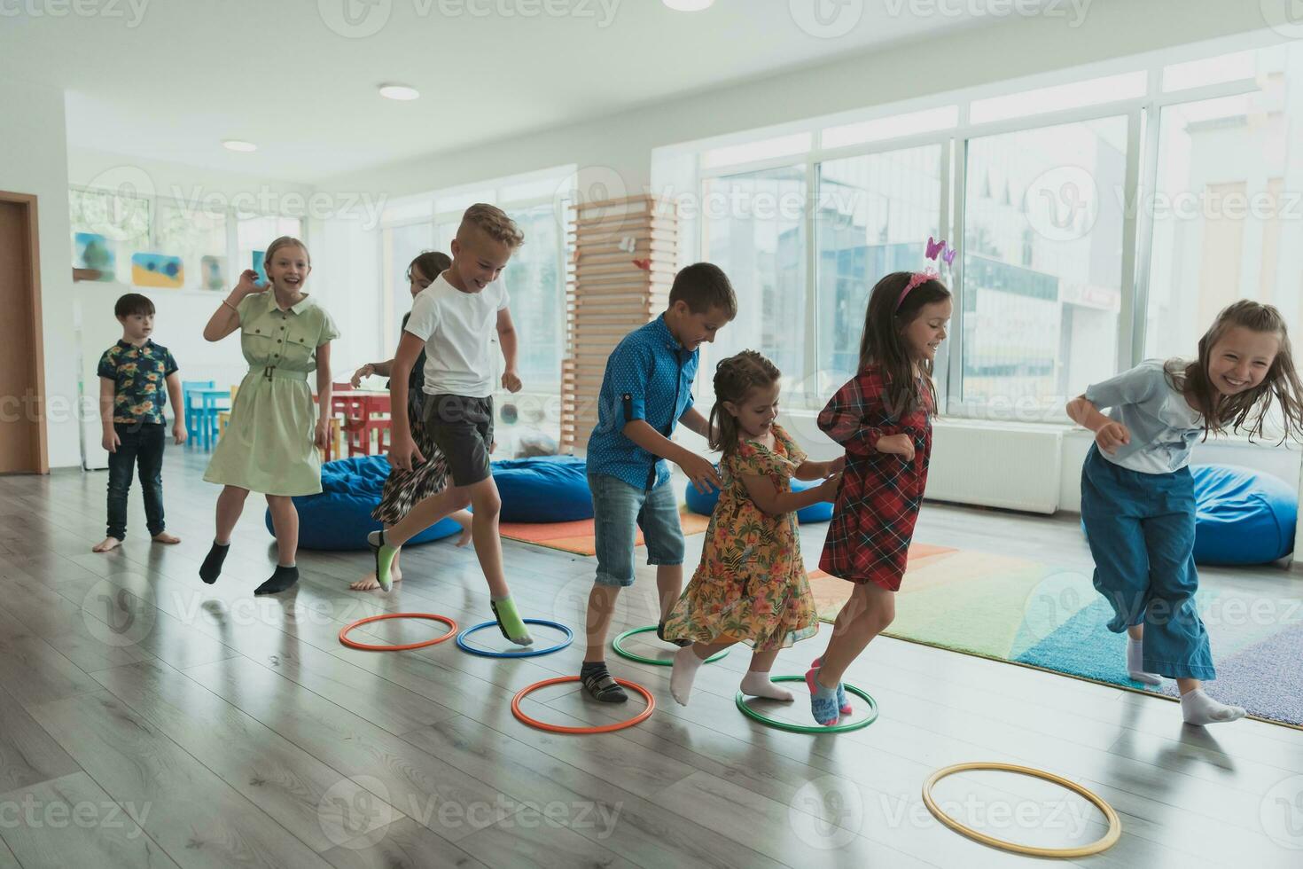 små barnkammare skola barn med kvinna lärare på golv inomhus i klassrum, håller på med träning. Hoppar över hula ring cirklar Spår på de golv. foto