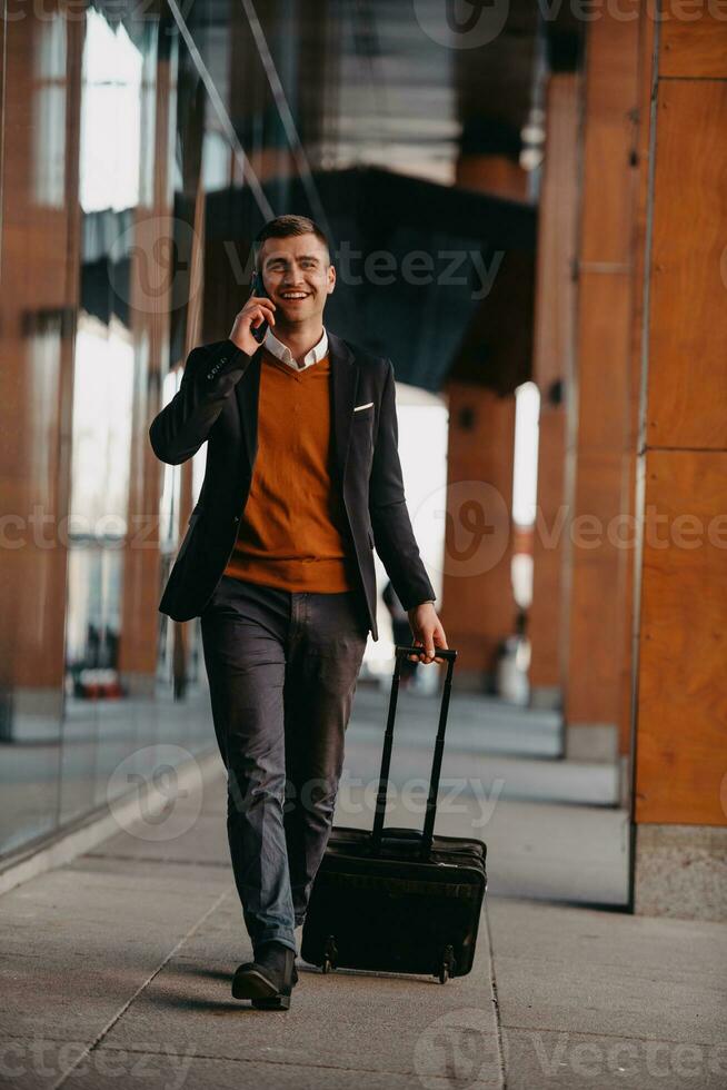 gående till flygplats terminal. självsäker affärsman resande gående på stad gator och dragande hans resväska dricka kaffe och tala på smartphone foto