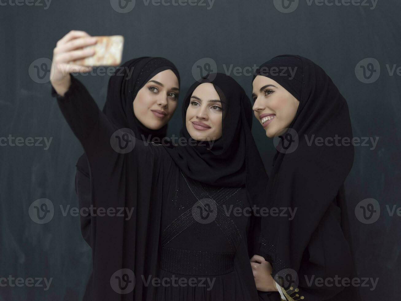 grupp av ung muslim kvinnor i modern klänning med hijab använder sig av smartphone medan tar selfie bild i främre av svart bakgrund foto