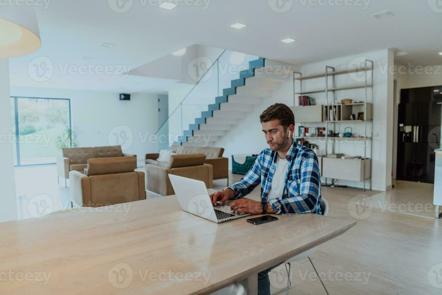 de man Sammanträde på en tabell i en modern levande rum, med hörlurar använder sig av en bärbar dator för företag video chatt, konversation med vänner och underhållning foto