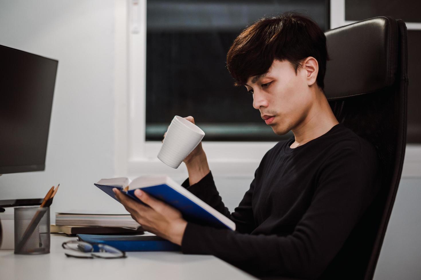 ung stilig asiatisk man som läser bok vid skrivbordet sent på kvällen foto