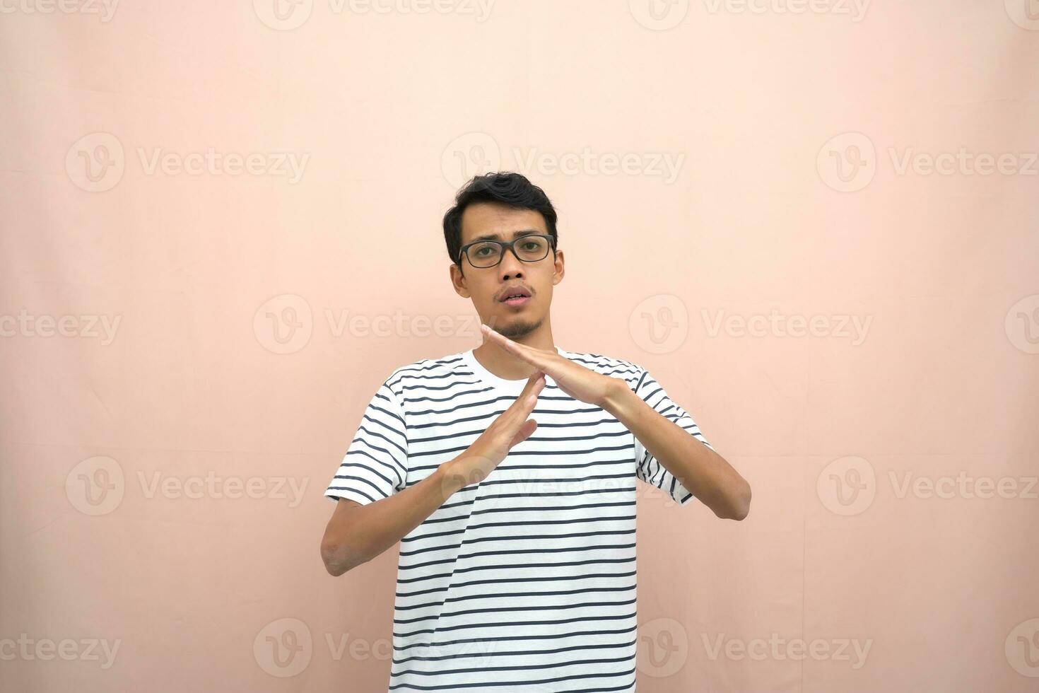 porträtt av ett asiatisk man i glasögon bär en tillfällig randig t-shirt. beteckna sluta, korsa förbud tecken, förbjuda något, vägra,. beige bakgrund. foto