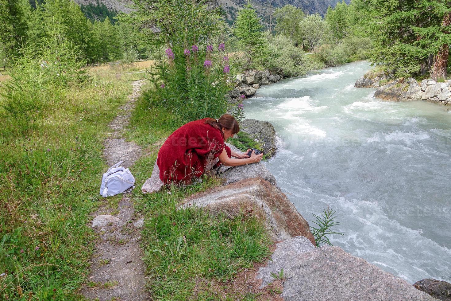 kvinna böjd ovanför bergsfloden som fotograferar landskapet foto
