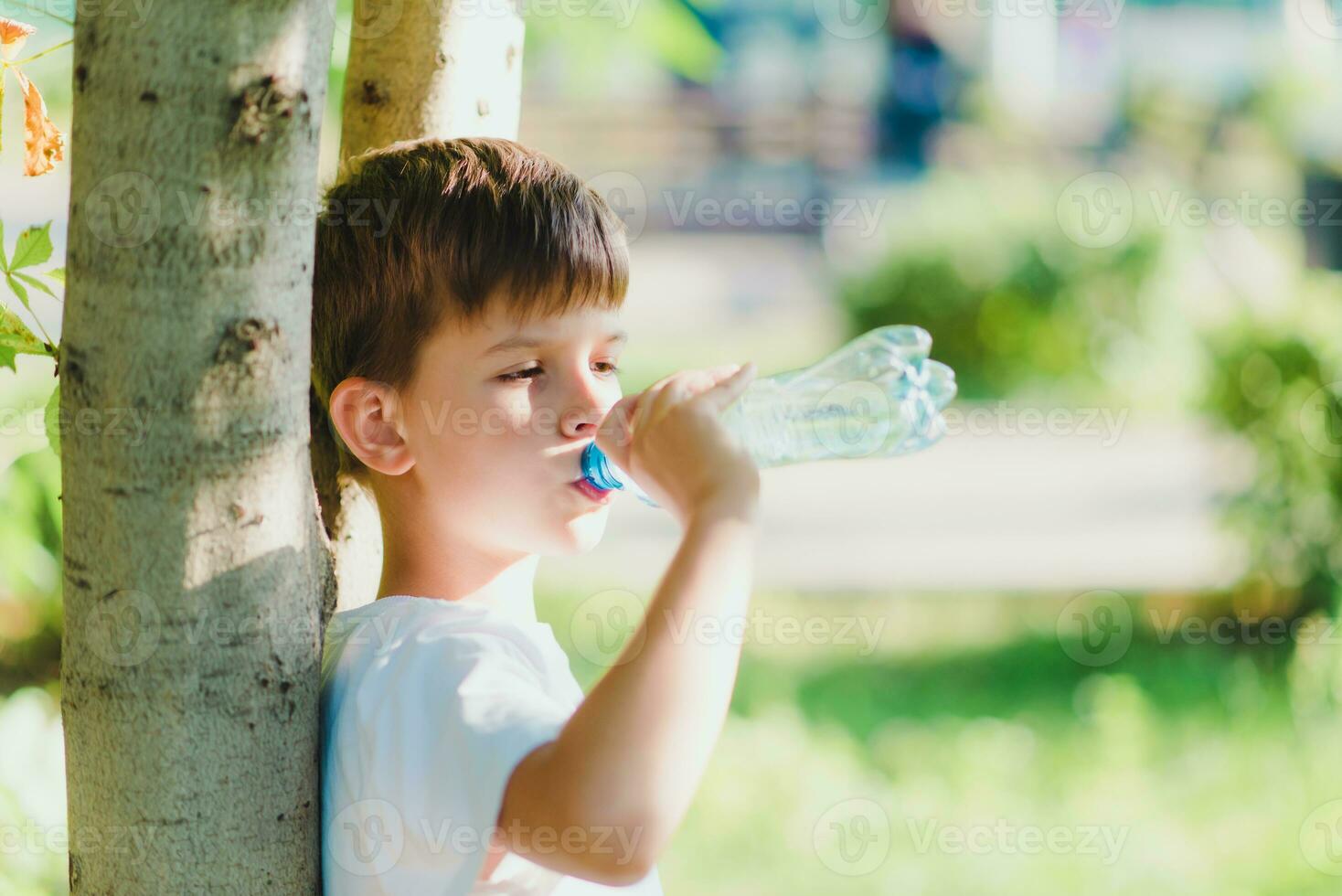söt pojke Sammanträde på de gräs drycker vatten från en flaska i de sommar på solnedgång. barn släcker törst på en varm dag foto